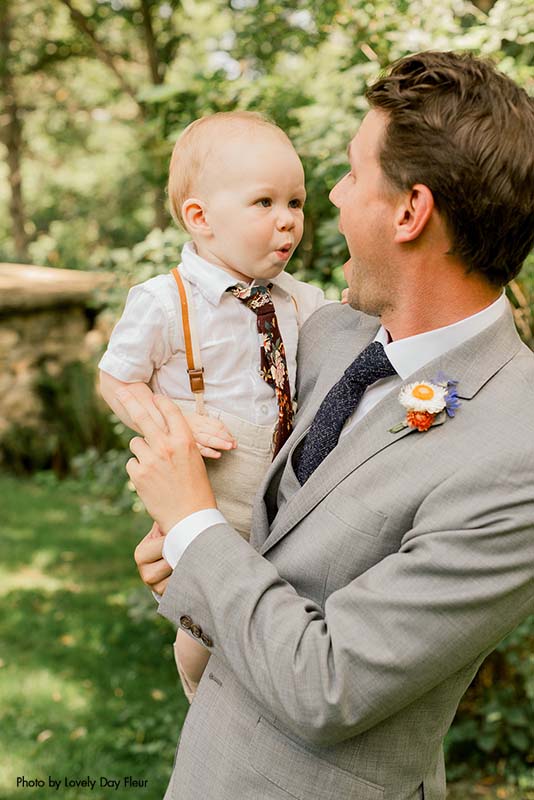 The groom in a gray suit holding a ring bearer in a white shirt and floral tie