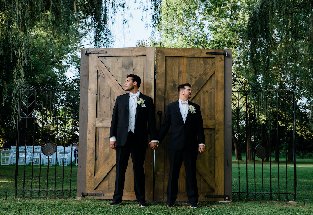 Modern outdoor wedding ceremony with two grooms standing in front of rustic barn door