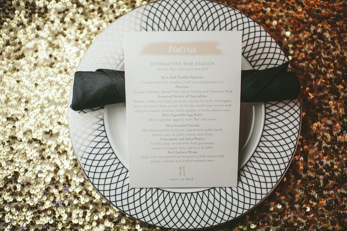 White wedding dinner menu sits on black rolled up napkin and white and black patterned plate