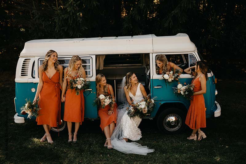 Bridesmaids in midi burnt orange dresses pose in blue Volkswagen van