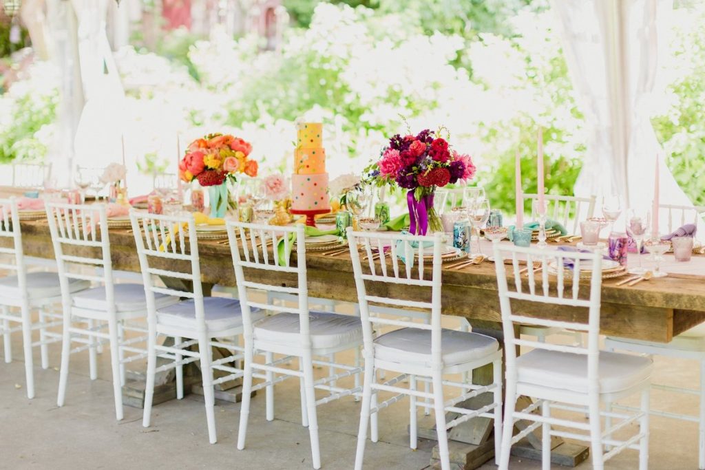 Bright spring wedding reception with white chairs and pastel-colored cake and flowers on top of a farm table