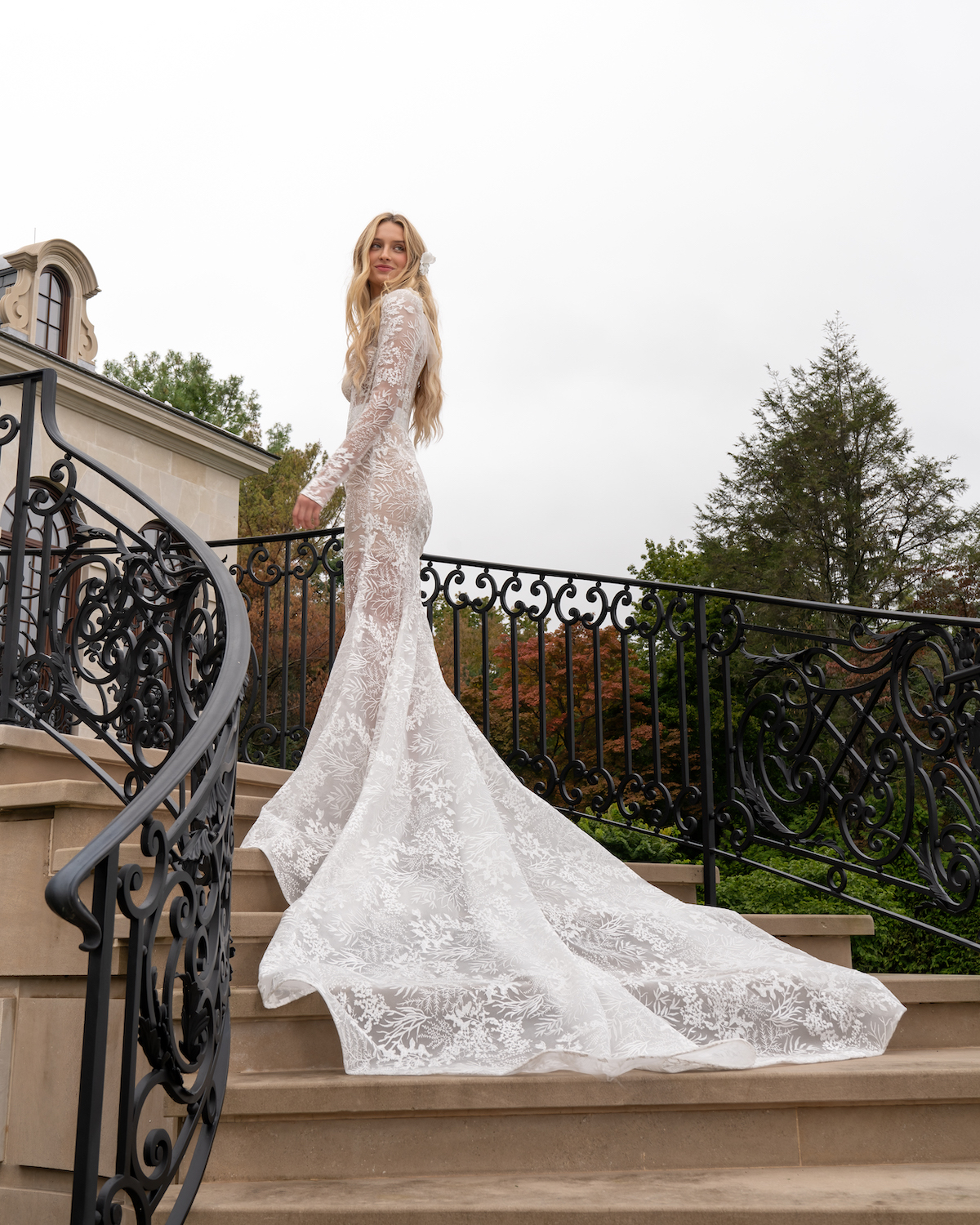 Bride stands on steps in trumpet bridal gown with lace sleeves by Reem Acra