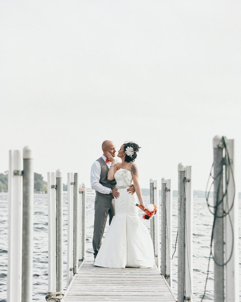 Bride and groom stand at the end of a dock on a lake by Zandolee Media