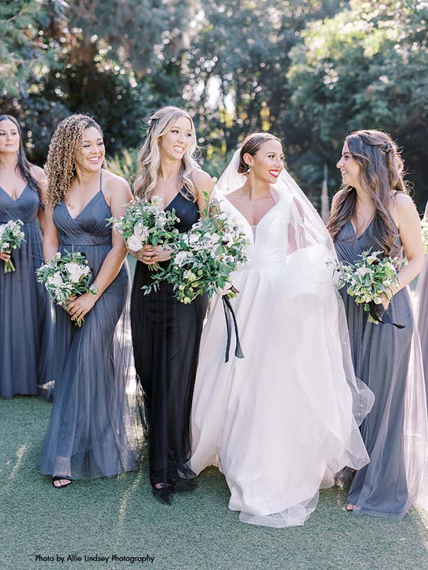 Bride in a-line v-neck satin gown walks through garden with bridesmaids in gray floor-length gowns