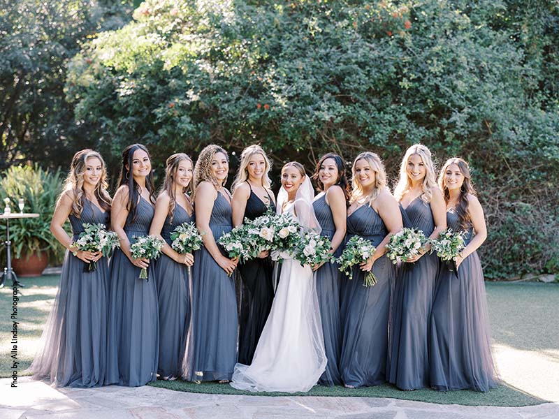 Bridesmaids in floor length blue-gray dresses stand with bride