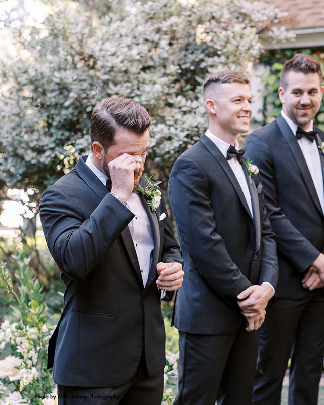 Groom in black suit wipes tear from his eye during wedding ceremony