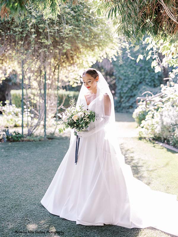 Bride in satin gown stands in garden with white and black bouquet