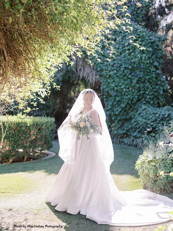 Bride in satin a-line gown stands in garden with veil draped over her face