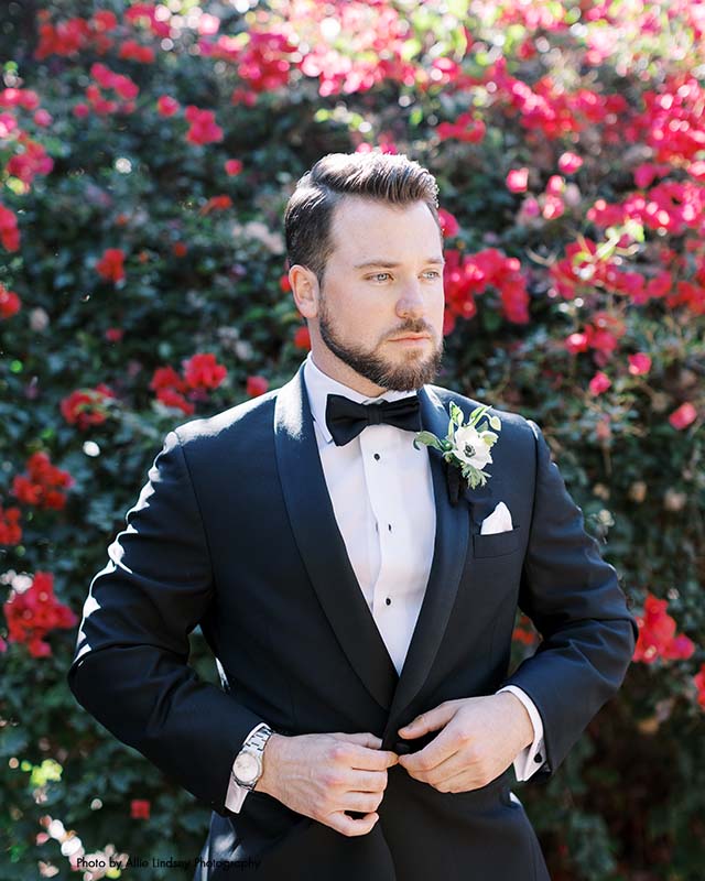Groom in classic black tux and bowtie stands in front of rose bush
