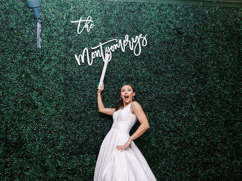 Bride stands in front of greenery hedge wall with neon sign