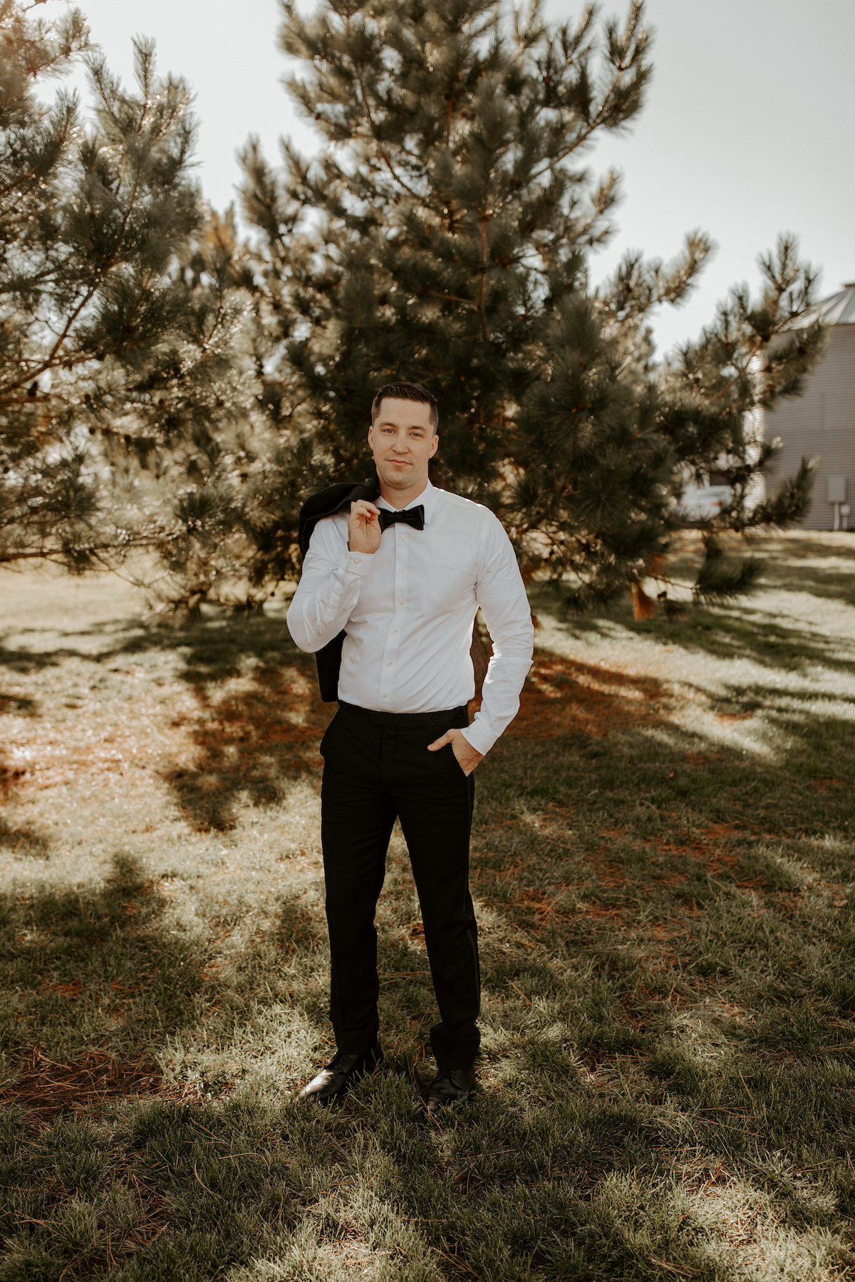 Groom in black suit and white shirt stands in front of fall trees