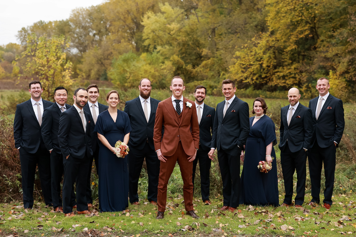 Groom in rust suit stands with wedding party in navy attire