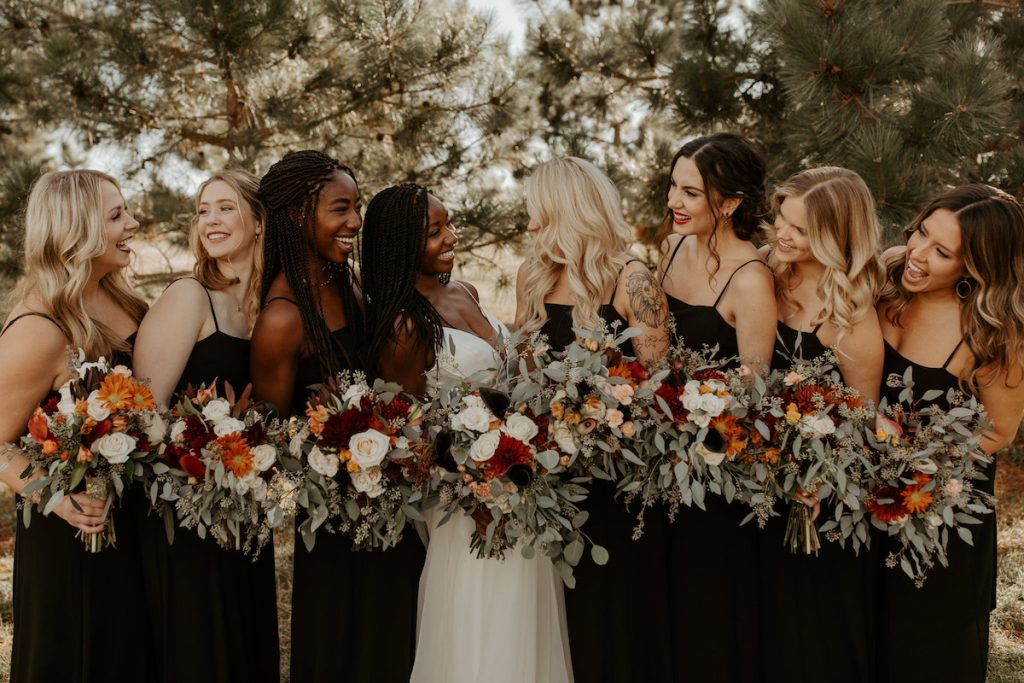 Bridesmaids in black dresses stand with bride with greenery, white, and red bouquets 